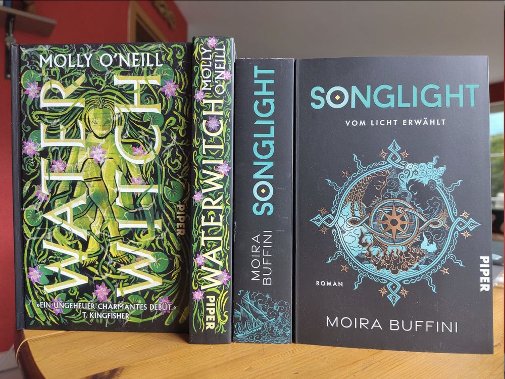 Die beiden Bücher Waterwitch (gebunden, grünes Cover mit einer Wassergestalt in einem Seeosenteich) und Songlight (Broschur, schwarzes Cover mit einem türkis-goldenen Emblem in der Mitte) stehen auf einem Tisch und sind von vorn und mit dem Buchrücken zu sehen. 