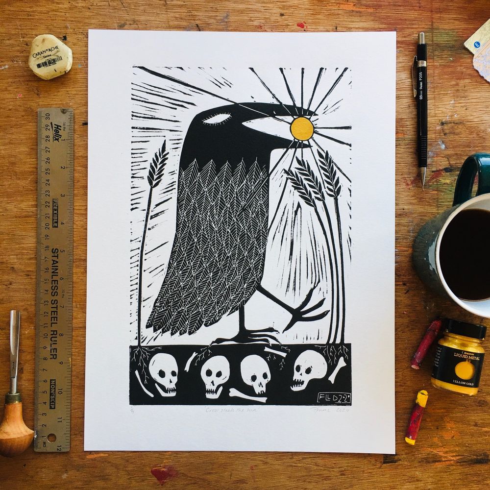 A lino print of a stylised crow walking among wheat. Below its feet are human skulls and bones. In its delighted beak is a gold shining sun.