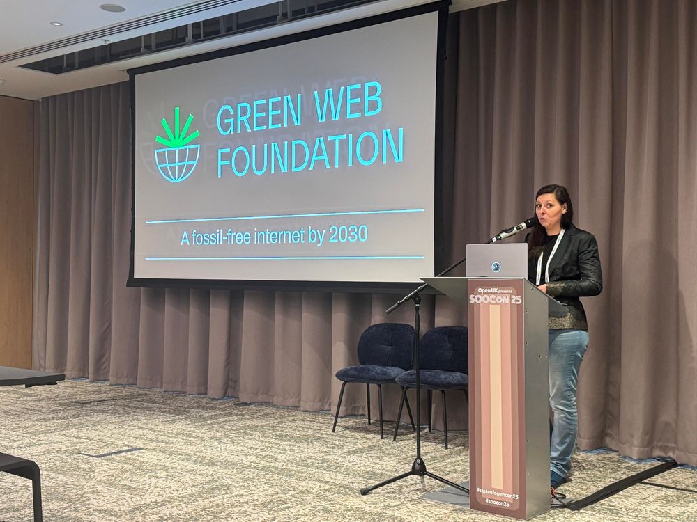 A slide with the a logo showing the lower part of a gridded globe with the upper half as a geometric green sprout, and the text GREEN WEB FOUNDATION A fossil-free internet by 2030. A woman with dark hair and wearing a dark jacket and blue jeans stands behind a podium with a microphone on a stand. 
