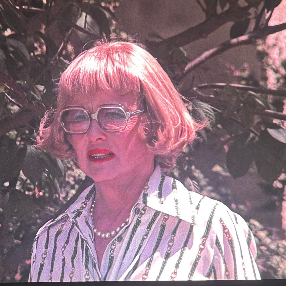 Bette Davis in Burnt Offerings. She has a kicky strawberry blonde bob and large glasses. She wears red lipstick, a pearl necklace, and a patterned shirt with a lapel that is wide by current standards but pretty subtle by 1976 standards. 