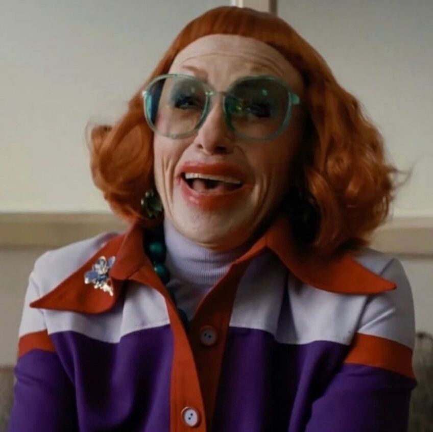 Amy Madigan in Weapons. She has a kicky red bob with incredibly short bangs and large glasses. She wears smeared red lipstick, a chunky necklace, and a lavender turtleneck underneath a colorblocked jacket with a lapel that is wide by both current and 1976 standards. 