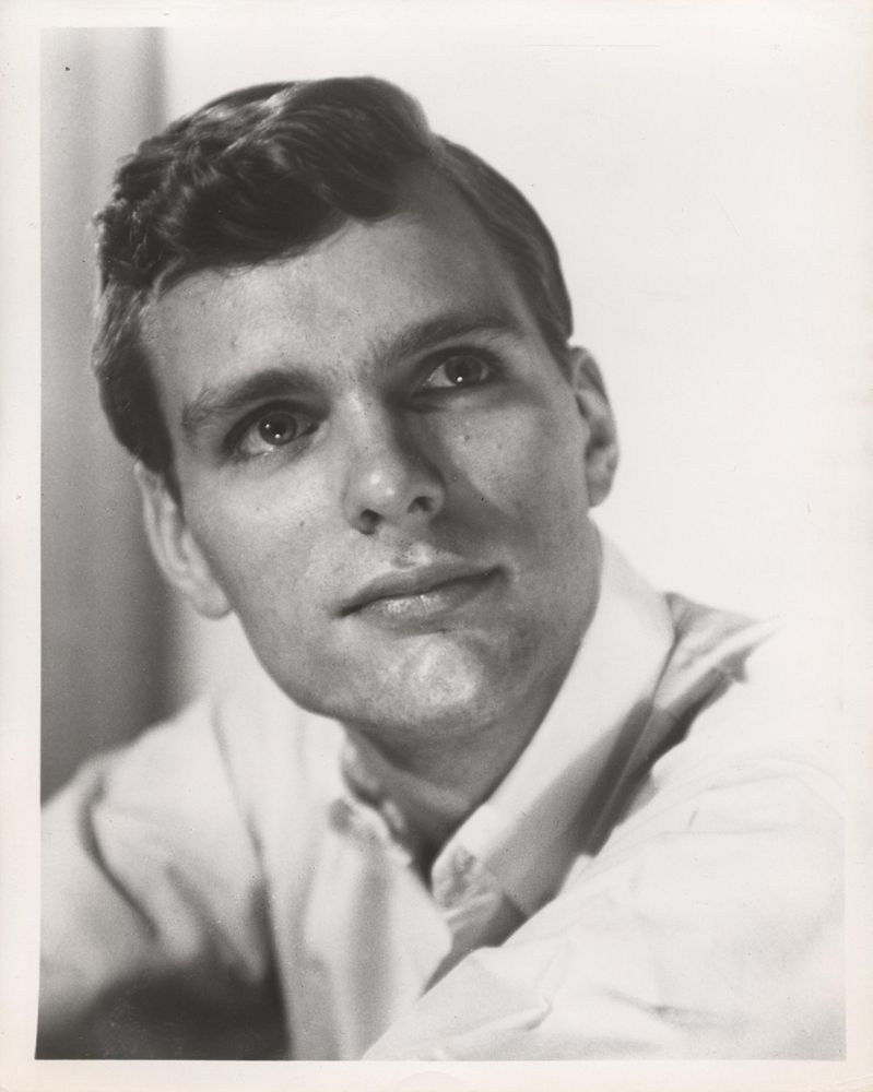 A black and white photo, presumably a headshot, of a young Keir Dullea. He is seen from the chest up and sits with his arms out in front of him. His body faces the left side of the photo and he turns his head to face the camera, looking up and to the right side of the photo. The man has a nice head of hair. 