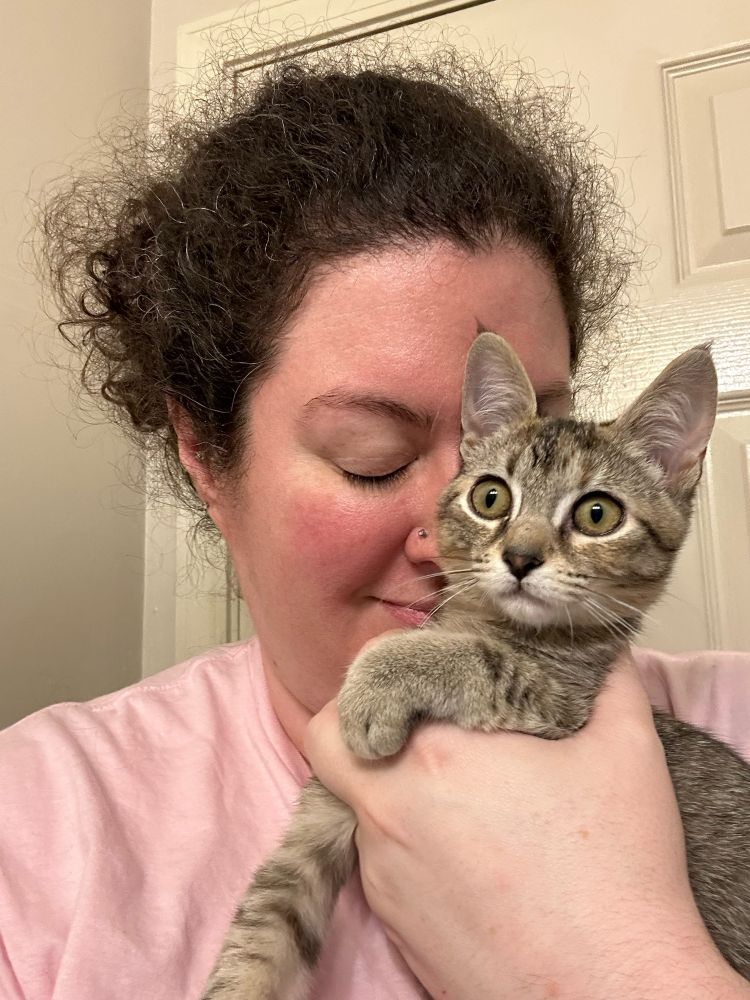 Jessica holds a tabby kitten, snuggling her with her eyes closed and a faint smile on her face. Tully the kitten looks into a mirror out of frame. Her eyes are wide as if she is seeing through space and time. 