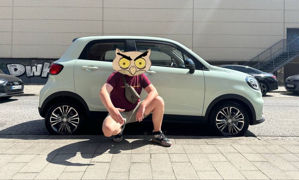 Person in kurzer Hose und Shirt kniet vor mintgrünem Kleinwagen an Straßenrand. Heinz-Eule auf Kopf montiert