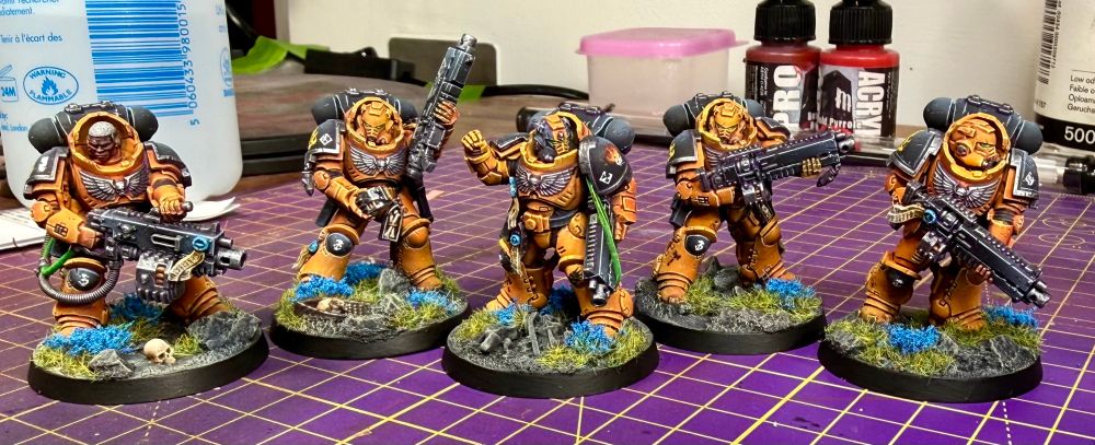 Five heavy intercessors of the Sons of Prometheus chapter, a successor chapter of the Salamanders. Their armour is orange with black pauldrons, backpacks and right knee pad. Their trim is dark gunmetal and weapons black, with blue purity seals and green eye lenses. Their bases are grey stone with green grass and blue flowers. Their company number is denoted by the ‘3’ on their right knee pads. The sergeant is marked out by a black helmet with orange vertical stripe. The marine armed with a heavy bolter is bareheaded, his skin is dark with grey hair and a stoic expression. 