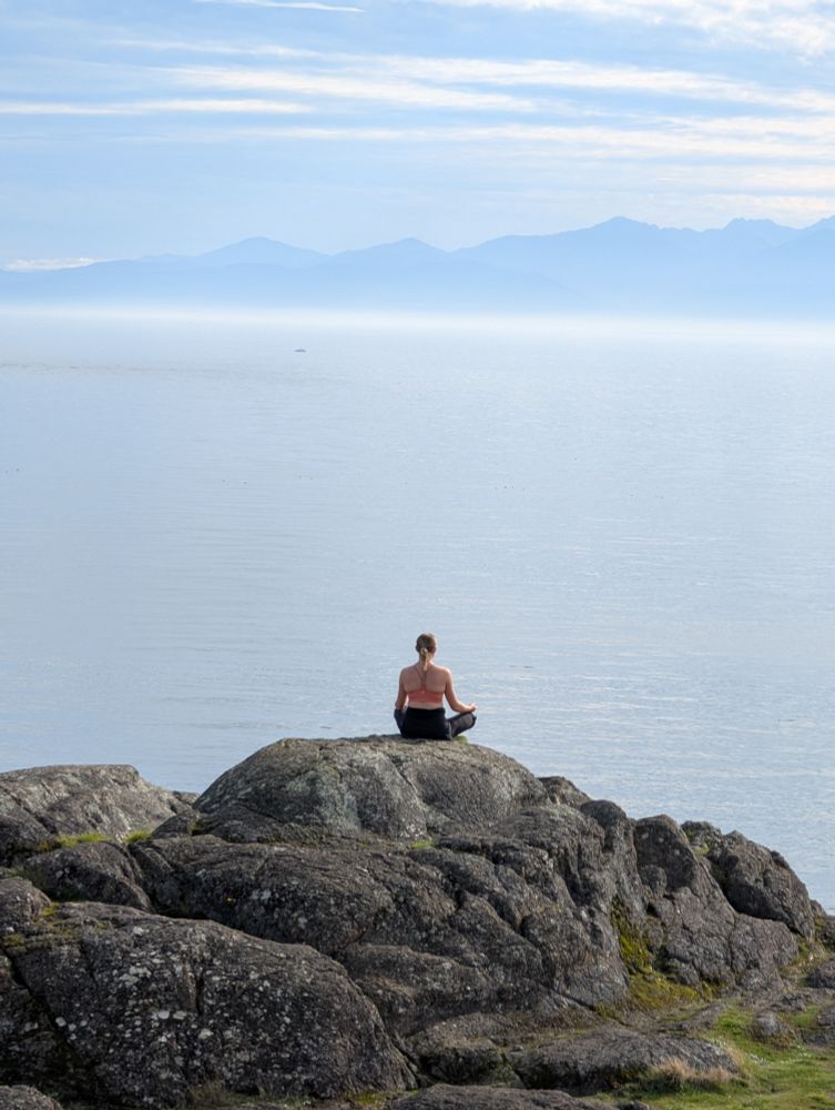A woman with her back to the camera, sitting on a rock in lotus pose, overlooking Salish Sea 