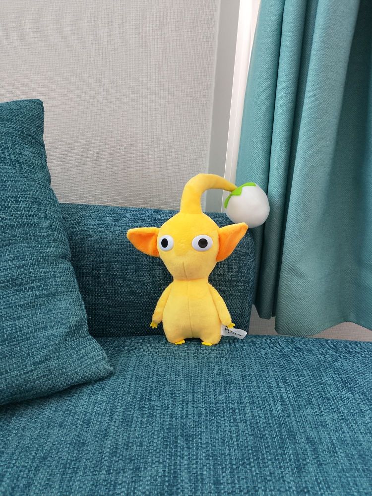 Yellow pikmin plushie sitting on a blue couch with light teal curtain in background