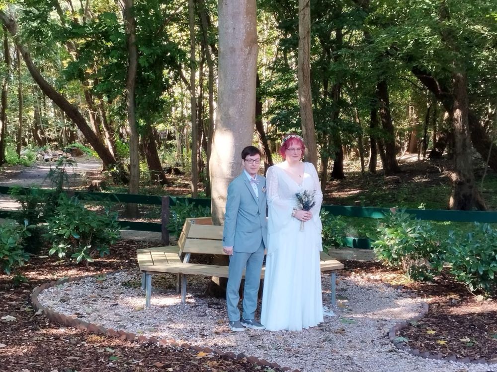 Mein Mann im Anzug und ich im Hochzeitskleid vor einem Baum um dem rundherum sitzbänke gezogen sind. Im Hintergrund ist Wald zu sehen. Ich halte noch einen brautstrauß mit beiden Händen vor mir.