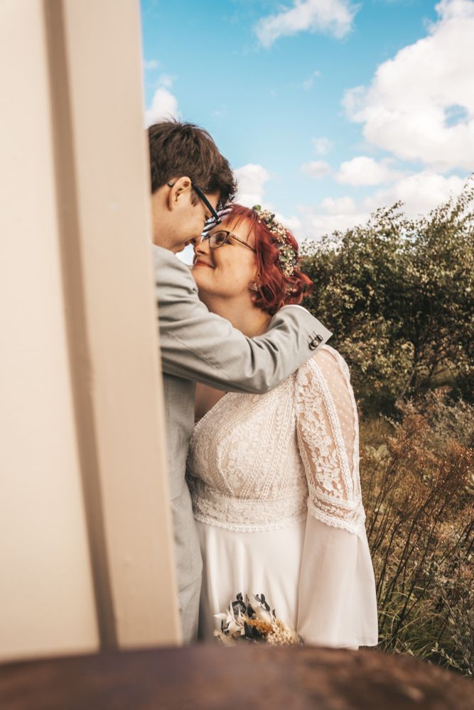 Man sieht vin der Seite über das Rad eines Badeskarren und an dessen Ecke vorbei auf uns. Im Hintergrund sind die Dünen. Nate hat die Arme um meinen Nacken gelegt und steht etwas erhöht. Er schaut zu mir runter, ich zu ihm hoch, nasenspitze an nasenspitze