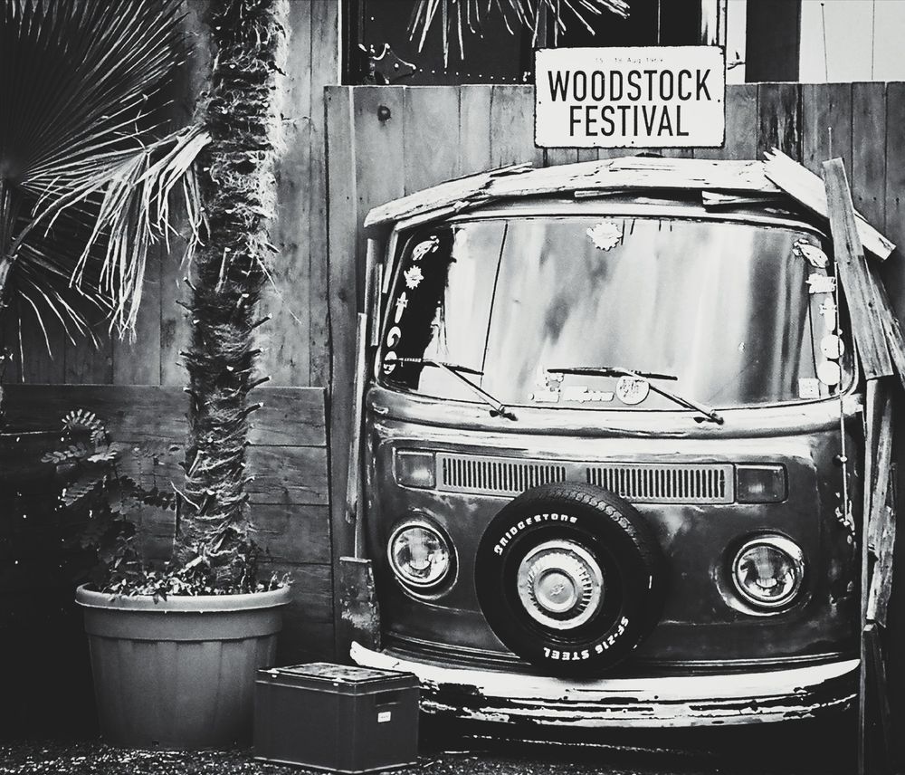 Ein schwarz-weißes Bild zeigt die Front eines alten VW-Busses, der in eine Holzwand integriert ist. Über dem Bus hängt ein Schild mit der Aufschrift "Woodstock Festival". Daneben stehen eine Palme in einem Topf und eine kleine Kiste.
