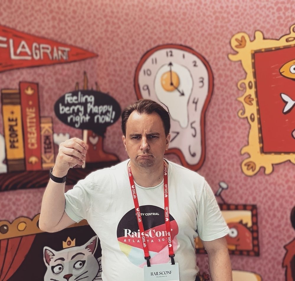A very serious looking Derek Houck holding up a chalk chat bubble sign that says, "Feeling berry happy right now!" against an abstract backdrop that feels like the fever dream of Salvador Dali if he watched too much Adult Swim.