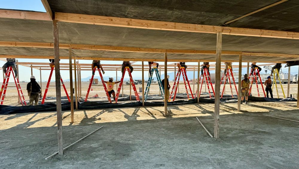 10 people on ladders, the upper half of their bodies obscured by a wood and cloth shade structure they are building/work on. 5 people stand on the ground as support for those on ladders.
