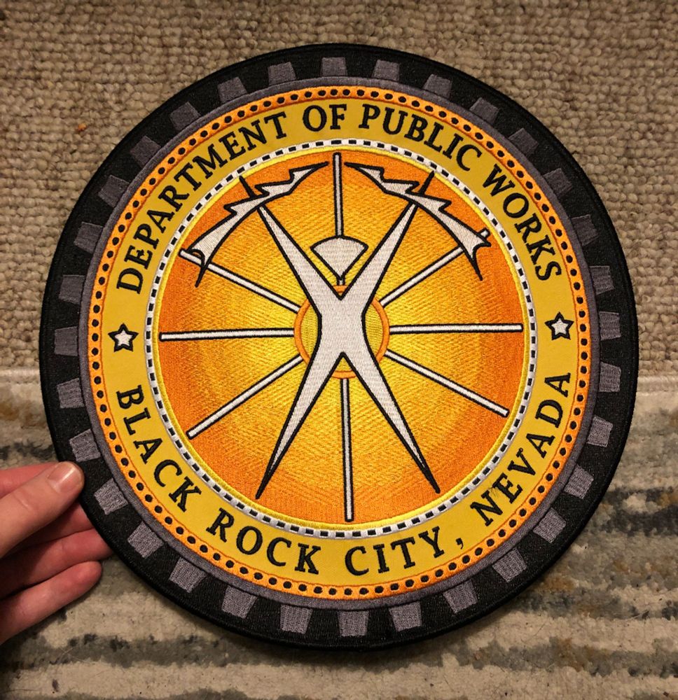 Circular 11 inch embroidered patch yellow to orange gradient from inside out with logo of Burning Man holding lightning bolts. Words embroidered "Department of Public Works, Black Rock City, Nevada." 