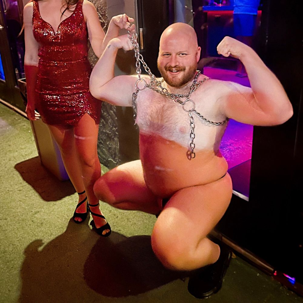 a woman in a red, sequin dress standing above a squatting & flexing bald, bearded man wearing a chain harness, a black leather thong & black combat boots