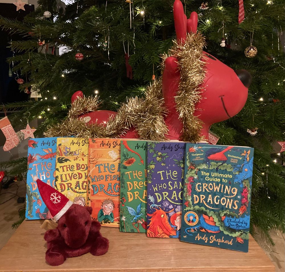 Photo shows all six books in The Boy Who Grew Dragons series written by Andy Shepherd and illustrated by Sara Ogilvie. In front of the books there is a small red cuddly dragon wearing a Santa hat and behind is an inflatable reindeer covered in a string of gold tinsel and a Xmas tree