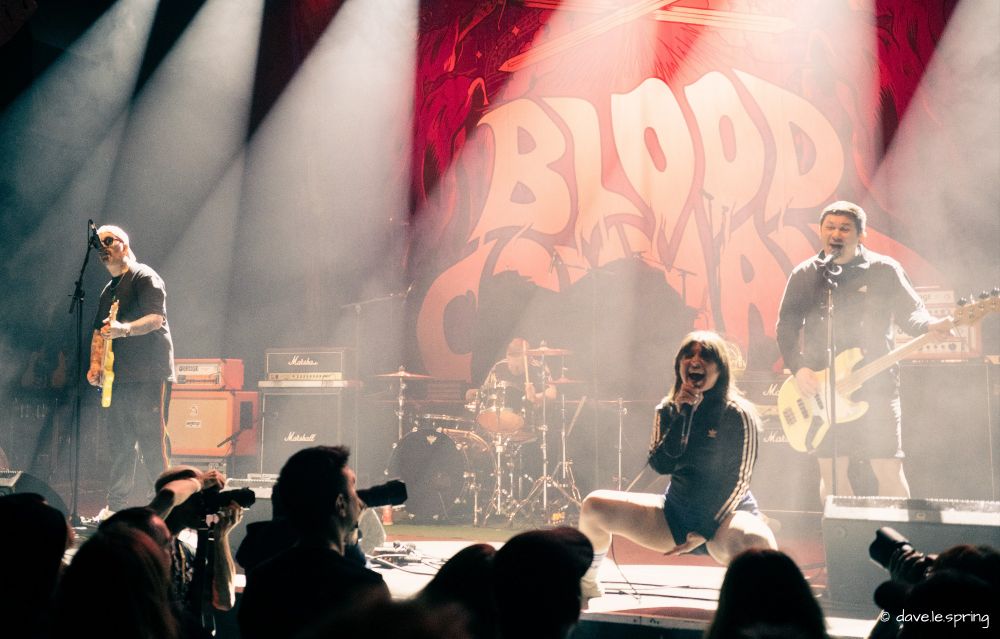 Picture of the band Blood Command in concert in Zurich on the 9.3.2025. it shows the whole band live on stage, rocking out during a song. There are multiple cameras pointing at the band from in front of the stage. Singer Nikki Brumen is squatting and screaming in her microphone while the rest of the band are playing their instruments and singing as well. The band logo can be seen behind the drum kit, there's a red hue on the picture but the bright stage lights throw white light everywhere.