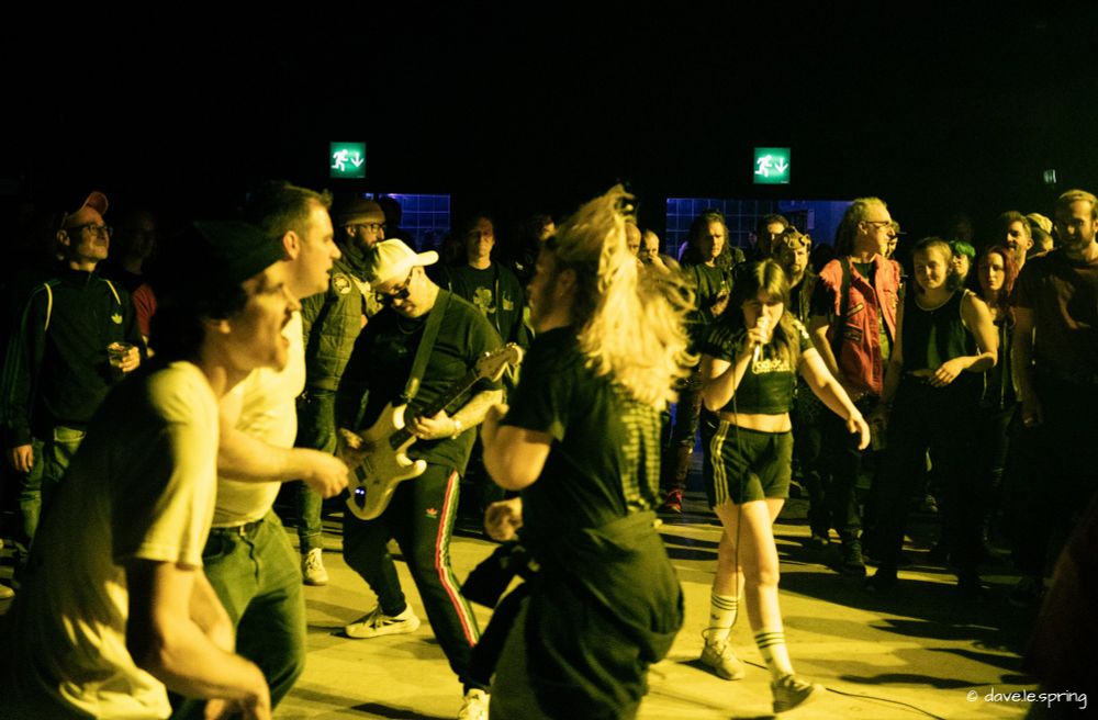 Picture of the band Blood Command in concert in Zurich on the 9.3.2025. it shows singer Nikki Brumen and guitarist Yngve Andersen in the middle of the audience. The picture is tinted in yellow light, members of the audience can be seen around the two musicians, Yngve is concentrating on playing the guitar while Nikki is looking at the camera and screaming in her microphone.
