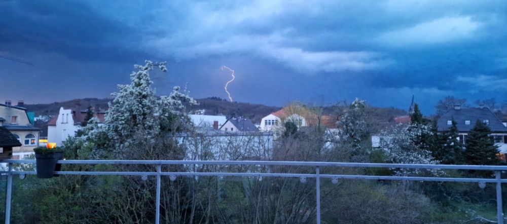 Ein dunkler Gewitterhimmel. Im Vordergrund des Bildes stehen Häuser. Im Hintergrund ist eine elektrische Entladung zu sehen.