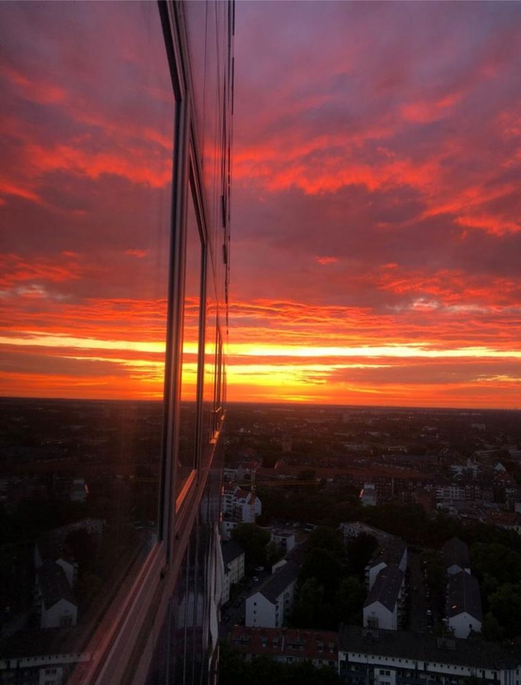 Blick aus einen Fenster eines Hochhauses in Köln. Zu sehen ist die Stadt unterhalb des Hauses, und der rote Abendhimmel. Die Stadthäuser wirken klein, und liegen im Dunklen. Der untergehende, rote Abendhimmel spiegelt sich in den geschlossenen Fenstern des Hochhauses wieder.