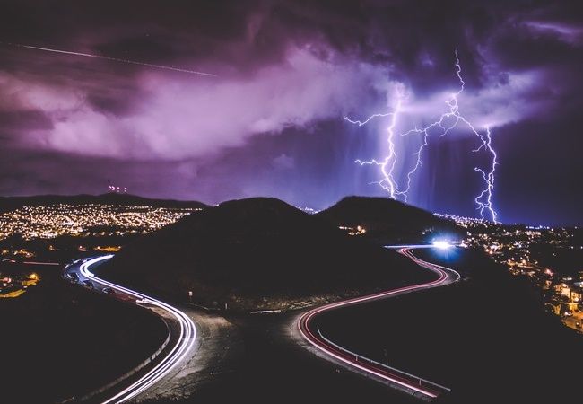 Purple night clouds and lightning above mountains and lit-up roads.
