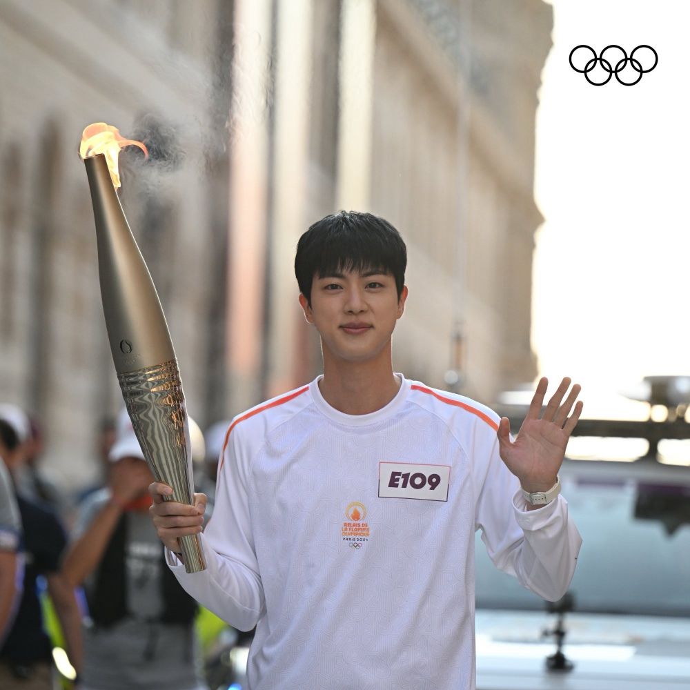 Kim Seokjin, popularly known as Jin, member of the group BTS, waves to the camera while participating in the 2024 Olympic torch relay. He is wearing the white uniform issued to all participants of the torch relay.