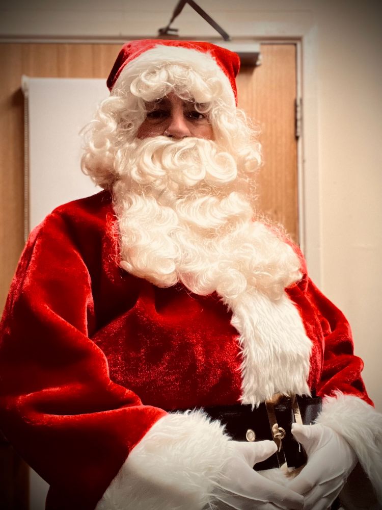 A photograph of a man posing in a father Christmas outfit with a large belly black belt, white gloves, red coat, and a big white beard and red hat