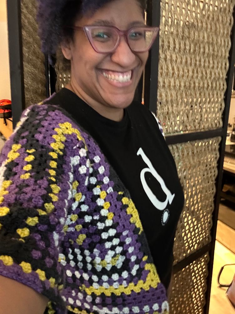 Kristen, smiling with teeth and wearing purple glasses, standing in front of a light wooden room divider. She has on a partially completed crochet hexagon sweater in the nonbinary colorway from JoAnn's house brand Big Twist.