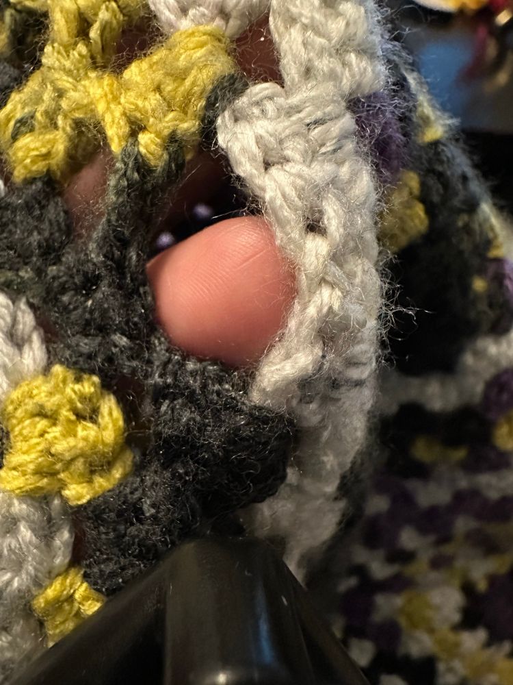 A closeup of the granny stitched black, purple and white fabric. Kristen’s finger is poking through a hole that shows a missed granny cluster stitch.