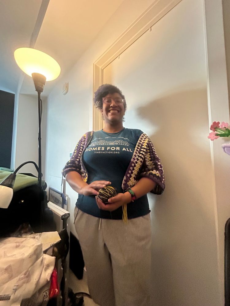 Kristen, a Black woman with a swooping Afro poof and purple glasses, a teal t-shirt that says Homes for All with a city skyline printed in white, under a purple, white, and black crochet granny sweater, stands and holds a ball of the yarn that makes up the sweater and looks directly at the camera. Her sweatpants are pants are grey 