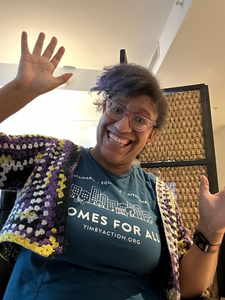Kristen, a Black woman with a swooping Afro poof and purple glasses, a teal t-shirt that says Homes for All with a city skyline printed in white, under a purple, white, and black crochet granny sweater, waves and smiles at the camera!