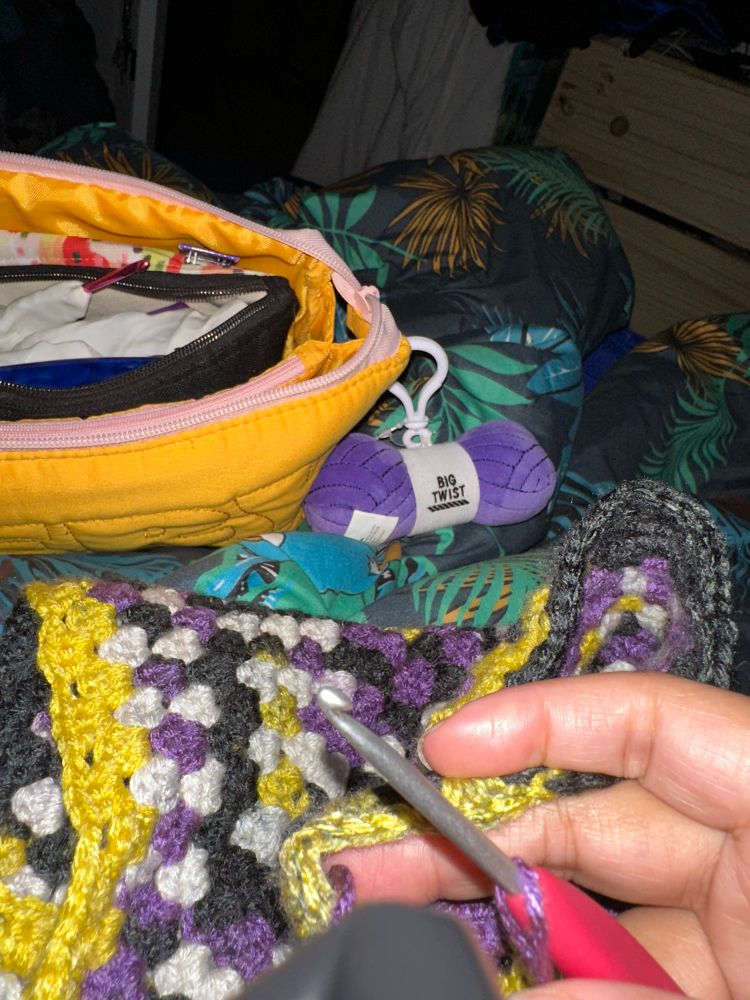 Kristen holds a Clover 3.5 mm crochet hook over her black, purple, yellow, and white crochet sweater. In the background is a golden yarn back with a purple plush yarn hank as a zipper pull. All of this is on a jungle print bed comforter.