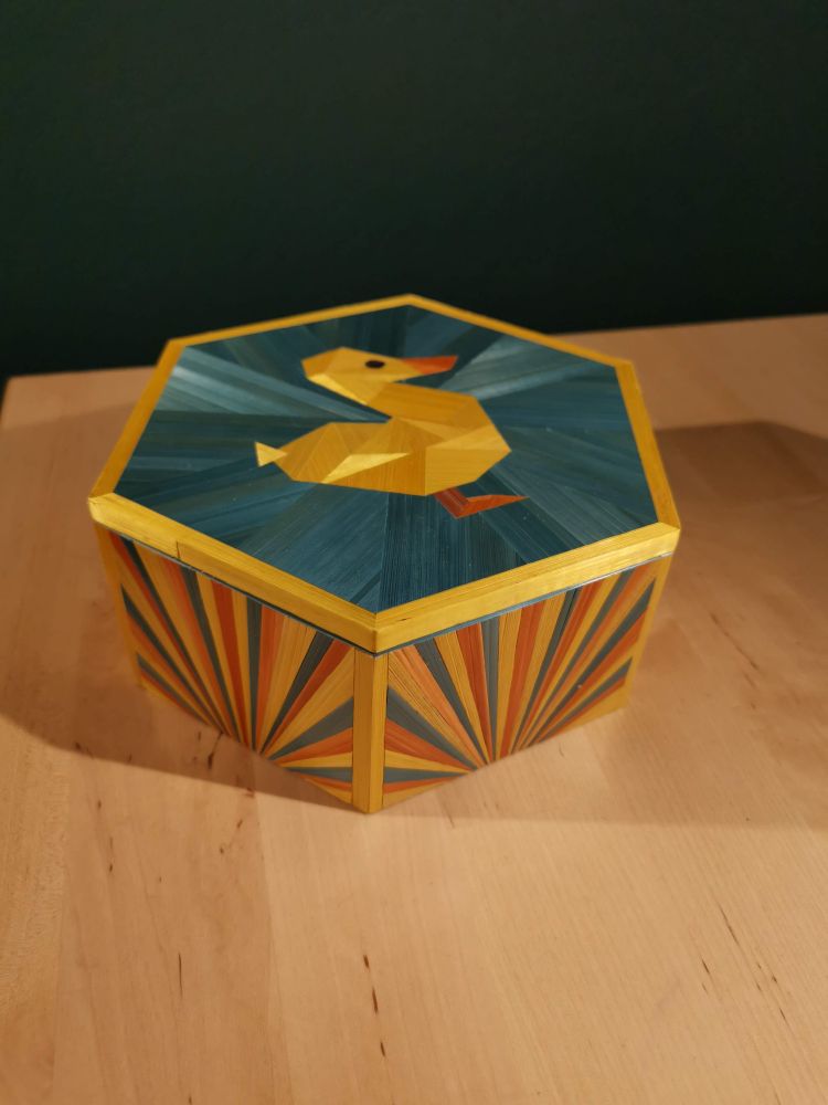 Boite hexagonale en marqueterie de paille, avec un caneton jaune façon origami, sur fond turquoise, sur le couvercle. Les côtés sont en motif soleil jaune, turquoise et orange 