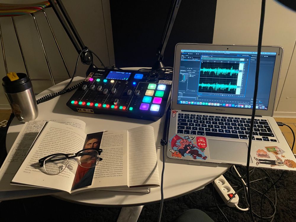 A photo of an audio recorder with a lot of bright colorful buttons and a laptop clearly being used to record audio, and an open book over some notebooks with messy notes scribbled 