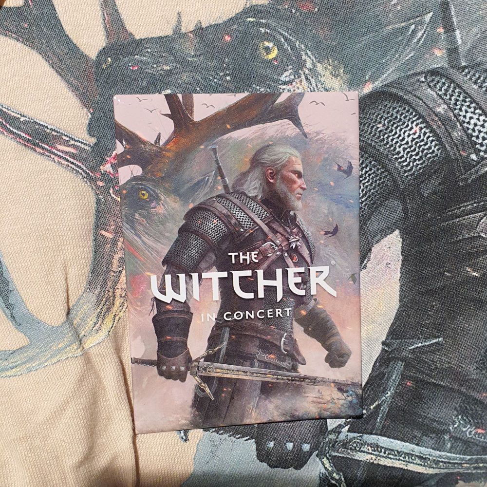 photo of a fridge magnet with geralt posed facing right and the words "the witcher in concert", sitting on top of a beige T-shirt with the same design.