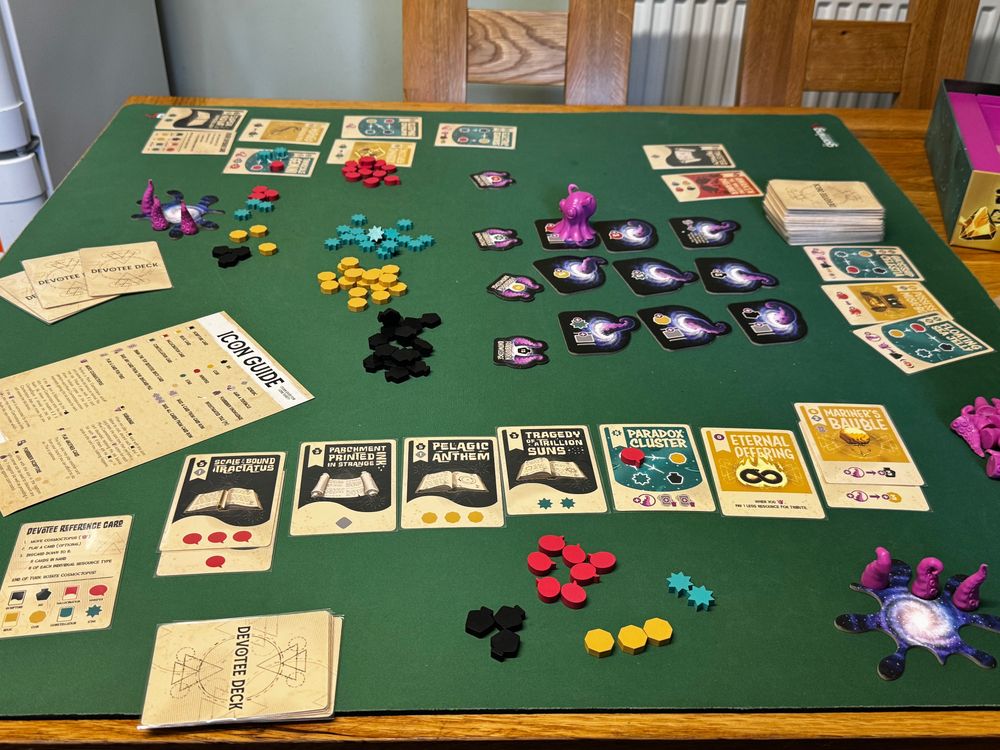Cosmoctopus board game setup on a green play mat showing various components including purple octopus miniatures, coloured tokens, different coloured cards and a grid of summoning locations in the middle.