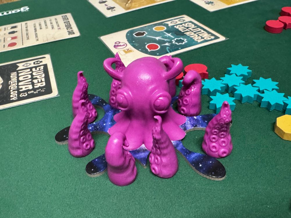 Close-up of a purple octopus miniature with eight tentacles arranged around it. The figure sits on a card base with a starry galaxy design, with inserts for each of its eight tentacles. Blue star-shaped tokens and game cards are visible in the background.