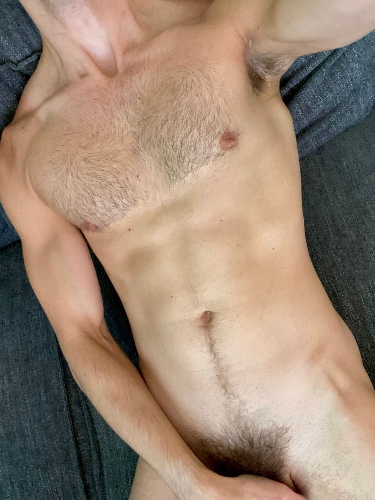 A naked man lying on a couch, one arm overhead to show pit while covering his cock with his hand and leaving his bush exposed. 