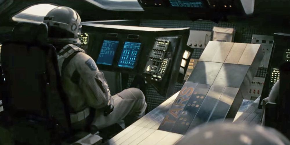 TARS, a robot resembling four rectangular metal bricks, in the cockpit of the Endurance from Interstellar. Two astronauts are present in the picture, both of their faces invisible. TARS' name is painted on TARS in both latin text and braille, and TARS is in the middle in between the two astronauts, in a pose resembling someone laying down.