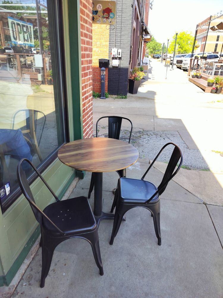 A modest wooden table with three metal chairs on a sidewalk outside the storefront 