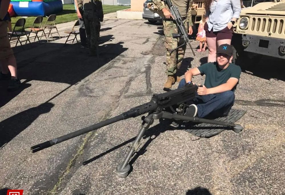 The white man who murdered Kirk is seated on the pavement with a gigantic gun that requires legs. Someone in camo with a rifle is behind him, as are several military Jeeps.