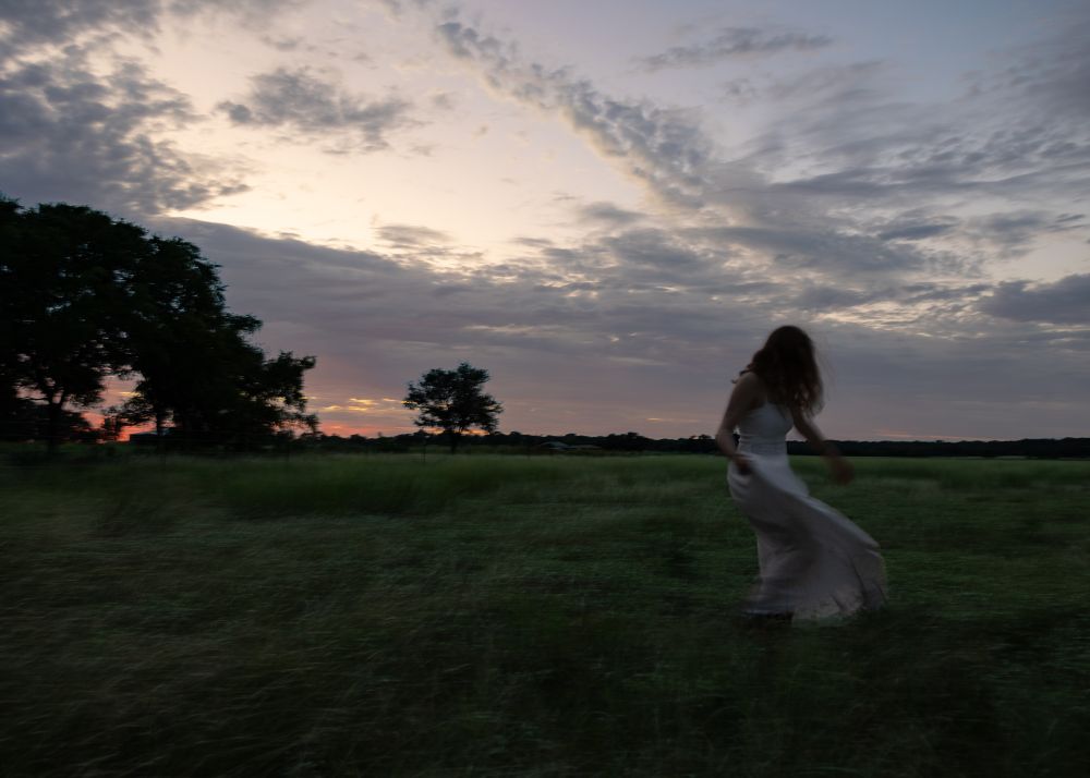 A woman in a white dress walks across a field at twilight. 