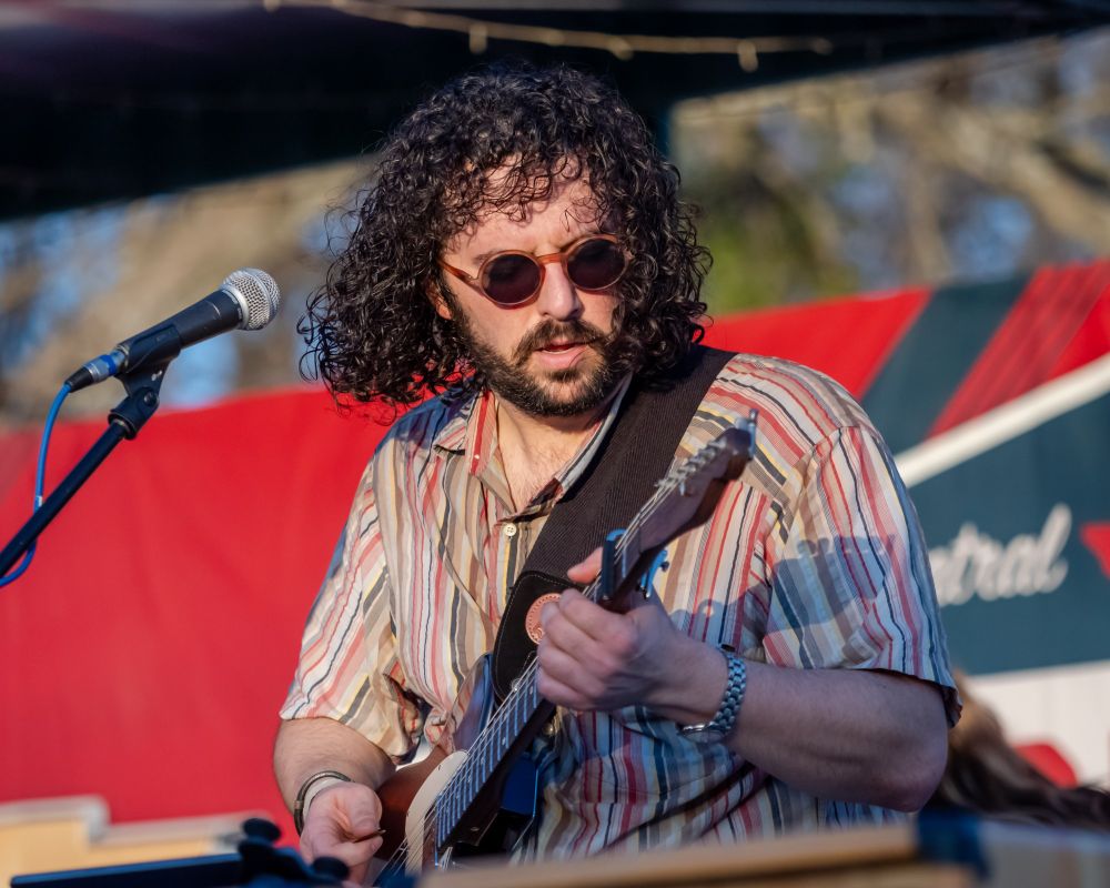 A guitarist with thick black curly hair and sunglasses playing outdoors during a festival. 