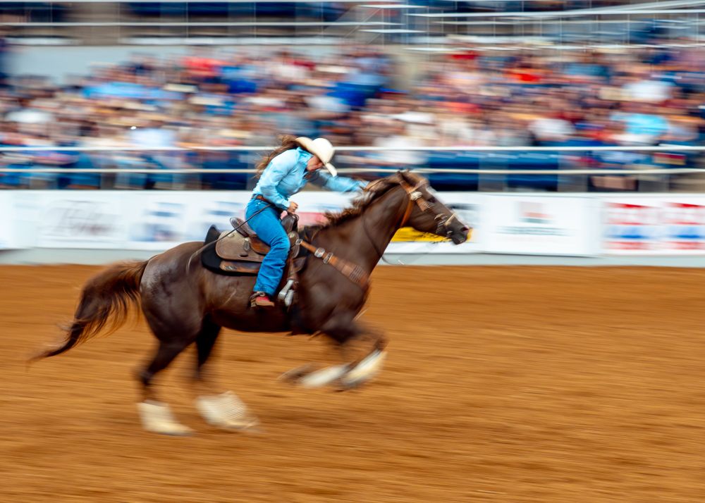 A cowgirl competing in the barrel racing at the rodeo. A slightly slow shutter speed creates a blurred effect. 
