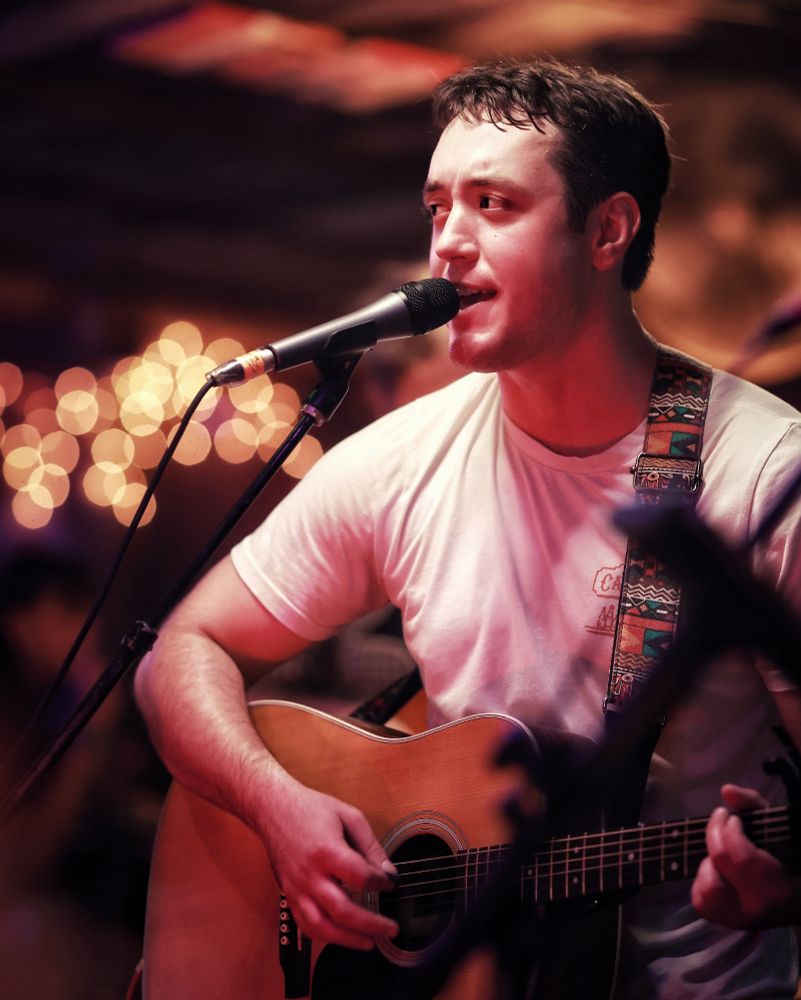 A young man playing guitar and singing in a honky tonk with an array of lights in the background. 