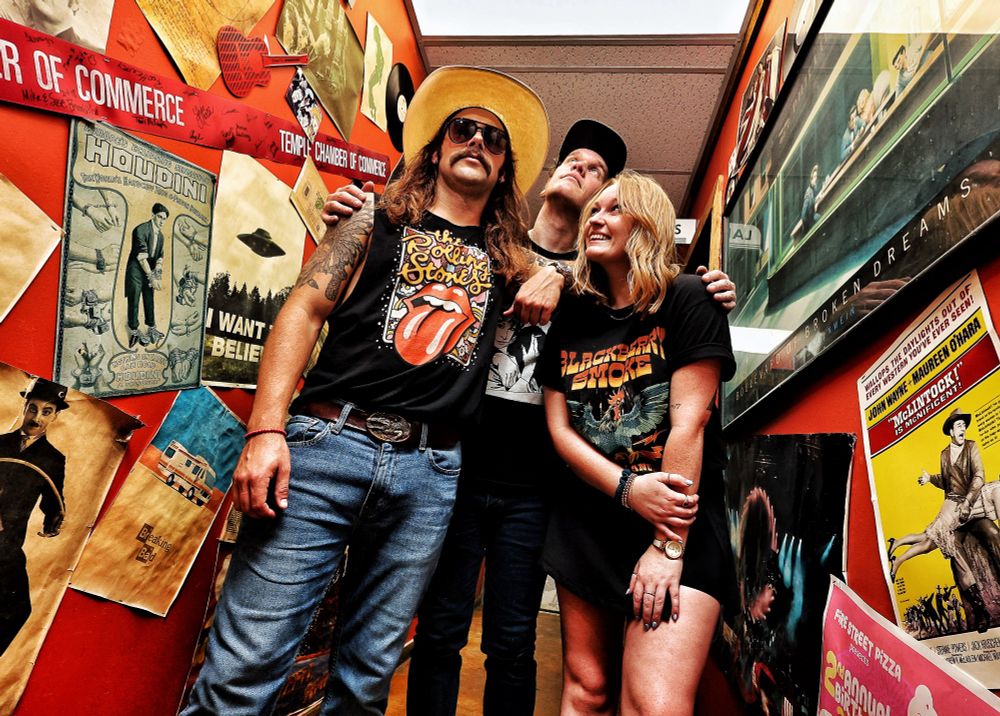 A three person rock band in a hallway decorated with a panoply of posters. 