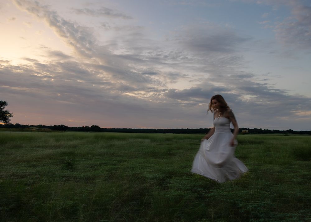 A woman in a white dress walks across a field at twilight. 