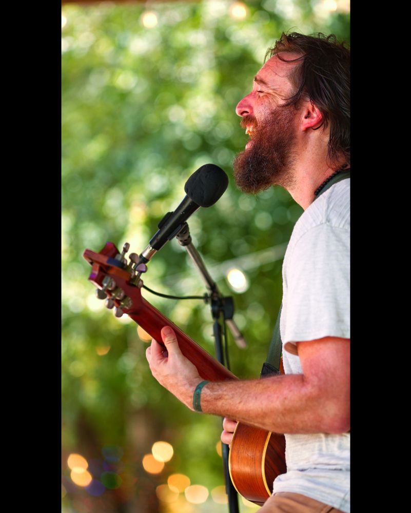 A bearded man singing and playing guitar with trees and lights in the background. 