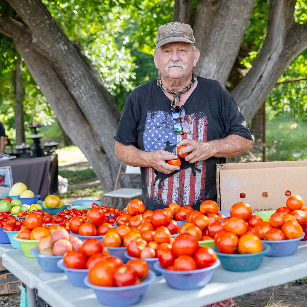 A farmer wearing a patriotic T-shirt and a camouflage hat stands in front of a table of fresh tomatoes for sale. 