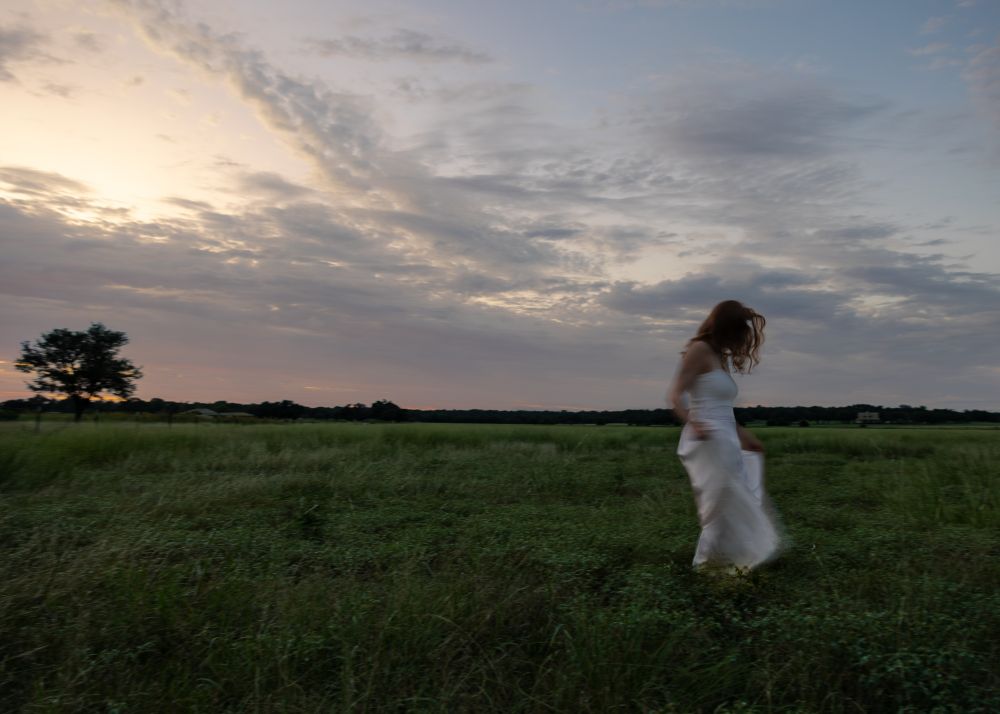 A woman in a white dress walks across a field at twilight. 