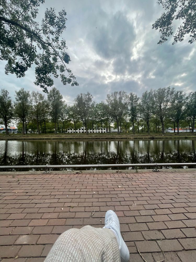 My leg in front of a red brick path in front of a stream with trees at the other side and a cloudy sky